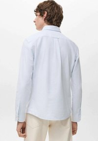 Joven con cabello corto castaño, que lleva una camisa de manga larga a rayas azul claro y pantalones color crema, de espaldas contra un fondo liso.