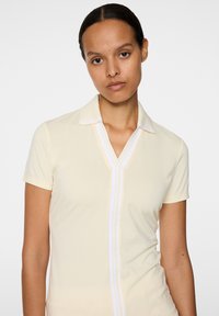 Żółta koszulka polo z krótkim rękawem, dekoltem w serek i białymi paskami wzdłuż listwy z guzikami. Gładka faktura, dopasowany krój.