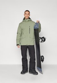 Zöld szigetelt dzseki cipzáras zsebből, fekete hó nadrág és fekete csizmák. A modell egy kék és fekete részletekkel ellátott snowboardot tart.