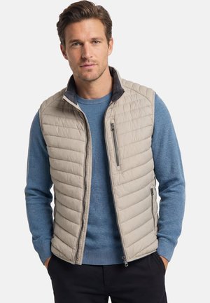 Homme portant une veste matelassée beige par-dessus un pull en maille bleu, les mains dans les poches d'un pantalon noir, debout devant un fond blanc.