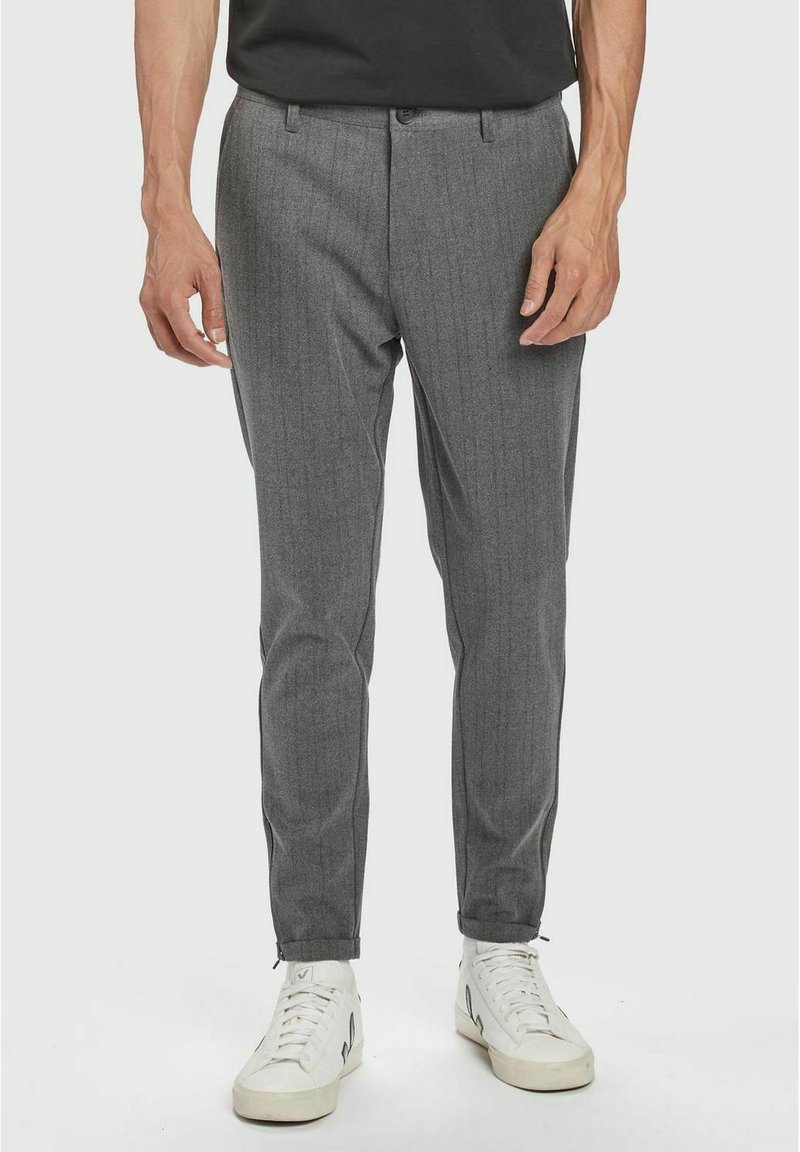 Graue, taillierte Hose mit vertikalen Nadelstreifen, schlanker Schnitt und umgenähten Säumen. Getragen mit weißen Sneakers vor einem schlichten Hintergrund.