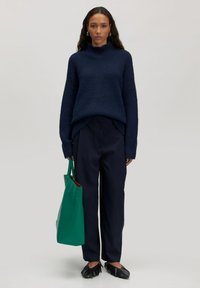 Marineblauer gerippter Pullover mit hohem Kragen, kombiniert mit weiten dunklen Hosen und einer grünen Tragetasche. Schwarze Loafers runden das Outfit ab.