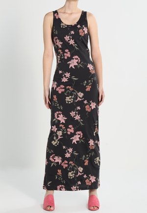 Robe maxi noire sans manches avec imprimé floral rose et beige portée avec des sandales ouvertes rose sur fond uni.