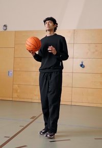 Sudadera negra y pantalones, sosteniendo una pelota de baloncesto naranja, de pie sobre un suelo de gimnasio con paredes de madera y un gancho azul visible en la pared.