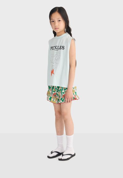 Jeune fille debout, portant un haut sans manches "PICKLES", un short à motifs colorés, des chaussettes blanches et des tongs noires sur un fond uni.