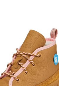 Botte enfant en cuir marron avec bordure rose, lacets rayés, visage de chat embossé sur la languette et étiquette circulaire bleue sur le côté.