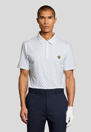Man draagt een wit poloshirt met korte mouwen en een blauw kruispatroon, donkerblauwe broek en witte golfhandschoen aan de rechterhand tegen een lichtgrijze achtergrond.
