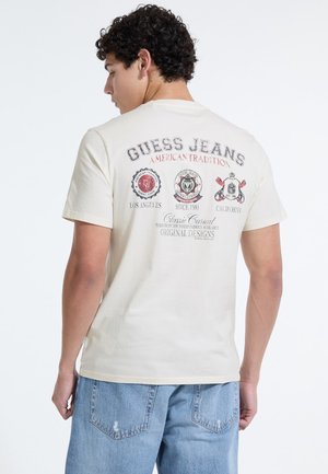 Lysbeige bomuld t-shirt med et grafisk print, der har cirkulære logoer og tekst på ryggen, parret med lyseblå denim jeans.