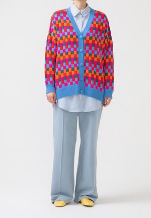 Personne porte un cardigan à carreaux coloré avec de gros boutons, une chemise rayée bleue, un pantalon large bleu clair et des chaussures jaunes, debout sur fond blanc.
