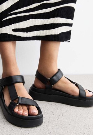 Sandali in pelle nera con cinturini regolabili, suola piatta e dettagli in fibbia. Soletta testurizzata visibile, abbinata a un indumento a righe bianche e nere.