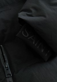 Svart pufferjacka med slät textur, utrustad med dragkedja och en svart etikett med präglad text. Lättviktsdesign och kompakt.