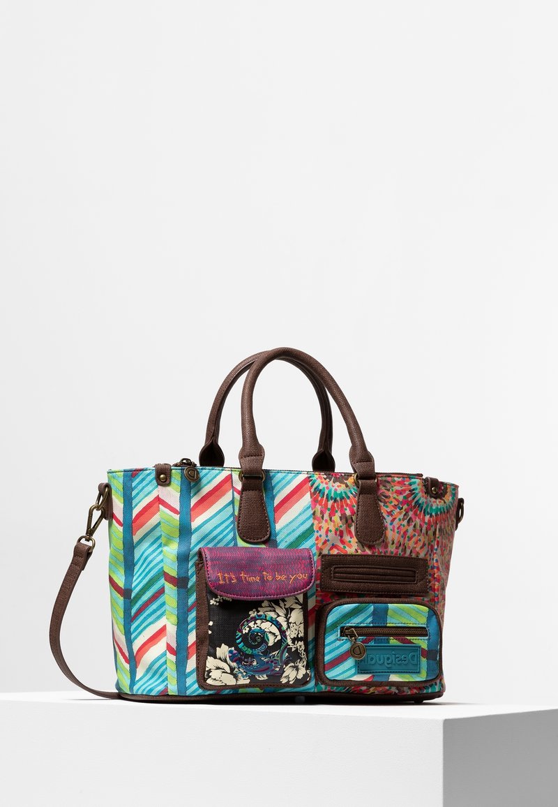 Sac à main coloré à motifs avec des poignées marron, deux poches avant et une bandoulière amovible sur une surface blanche sur un fond uni.