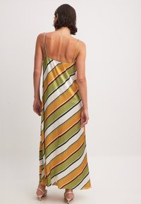 Robe maxi à fines bretelles, présentant des rayures diagonales en jaune, vert et blanc. Texture soyeuse avec une coupe décontractée et un dos ouvert.