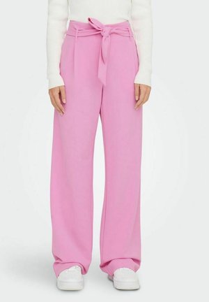 Person trägt hoch taillierte, weit geschnittene rosa Hosen mit vorderer Schleife, kombiniert mit weißen Turnschuhen und einem weißen Langarmshirt.