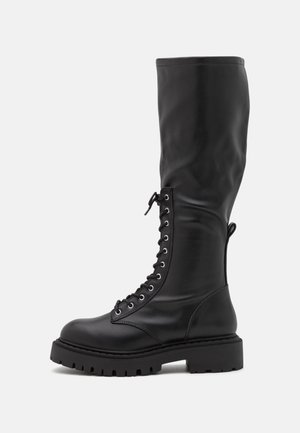Botte haute en cuir noir à lacets avec semelle épaisse à crampons et languette à l'arrière, présentée sur un fond clair uni.