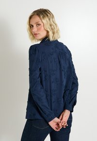 Camisa de manga larga azul marino con bordado floral, puños de botón y un corte holgado. Textura suave con un diseño de cuello clásico.