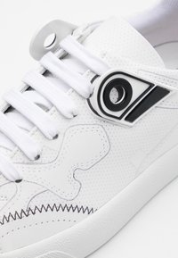 Biały sneaker z perforowaną powierzchnią, z czarnymi i szarymi akcentami. Zawiera białe sznurowadła oraz unikalny czarno-biały detal logo.