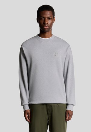 Grå sweatshirt med långa ärmar och rund halsringning, med en liten broderad logotyp på bröstet, i kombination med olivgröna byxor.