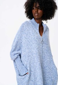 Blauwe gebreide trui met een hoge kraag, volledige rits en geribde manchetten. Textuurpatroon over de hele trui. Voorzien van zijzakken voor extra functionaliteit.