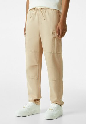 Beige cargo pants with an elastic waistband and drawstring. Features side pockets, a relaxed fit, and stitched panel details.