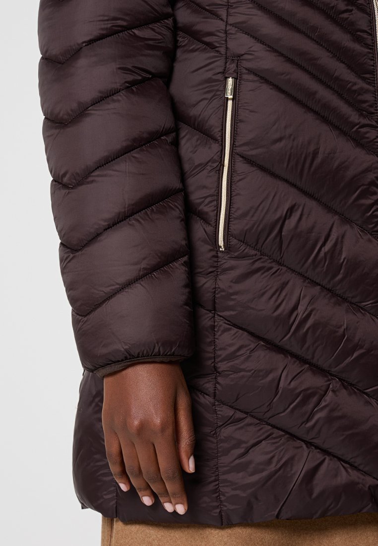 Brązowa kurtka puchowa z diagonalnym wzorem pikowania, metalowymi zamkami błyskawicznymi oraz gładką, błyszczącą teksturą. Zbliżenie na rękaw i dłoń.