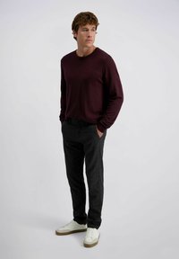 Bordeaux lange mouwen trui gepaard met donkere gestructureerde broek en witte sneakers met beige accenten; staande houding tegen een eenvoudige achtergrond.