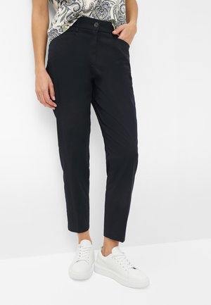 Persona che indossa pantaloni neri a sigaretta, sneaker bianche e un top fantasia, in piedi con una mano in tasca su sfondo bianco.