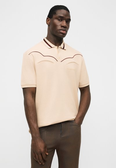 Άνδρας που φοράει μπεζ κοντομάνικο πόλο πουκάμισο με σκουρόκαφέ περίγραμμα, στέκεται με το ένα χέρι στην τσέπη μπροστά σε απλό φόντο.