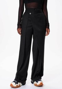 Zwarte wijde broek van soepel materiaal, met een hoge taille en een diagonale knoopsluiting, alsook met plooien aan de voorkant. Gestyled met sportieve sneakers.