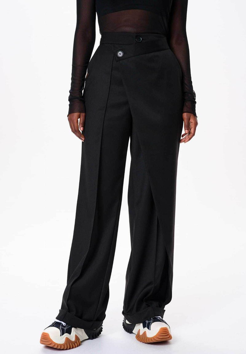 Zwarte wijde broek van soepel materiaal, met een hoge taille en een diagonale knoopsluiting, alsook met plooien aan de voorkant. Gestyled met sportieve sneakers.