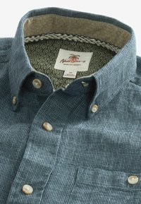 Camisa de botones en un tono azul verdoso oscuro con un patrón de cuadros. Presenta botones crema, estampado en el cuello y un bolsillo en el lado izquierdo. Etiqueta visible en el interior.