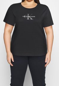 Camiseta de algodón negra con cuello redondo, mangas cortas y un logo gráfico plateado "Calvin Klein Jeans" en el frente.