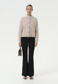 Beige gebreide cardigan met een hoge kraag en zwarte knopen, gecombineerd met zwarte flare jeans en een kleine zwarte handtas.