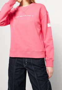 Felpa rosa con una vestibilità comoda, presenta un collo a V e polsini a coste. La scritta recita "Perché non c'è un pianeta B." Abbinata a pantaloni in denim scuro.
