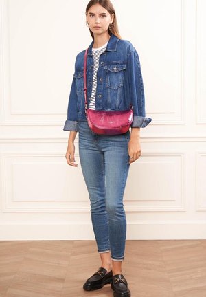 Sac en bandoulière métallisé rose, de forme incurvée, porté avec une veste en denim, une chemise rayée et un jean skinny sur un fond neutre.