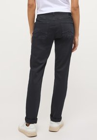 Egy személy háttal áll a néző felé, sötét farmert, fehér pólót és fehér sportcipőt visel, világos háttér előtt.