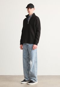 Crna zip-up dukserica, svijetloplave traperice i crne tenisice. Model nosi crnu kapu, minimalistički dizajn, glatka tekstura, opušten kroj.