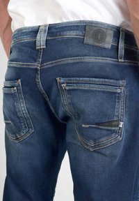 jeans de mezclilla con un lavado azul oscuro, diseño estándar de cinco bolsillos, costuras visibles y un pequeño parche de cuero en la cintura.