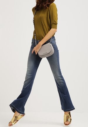 Vrouw die een mosterdkleurige trui, wijdlopende blauwe jeans, gouden sandalen en een kleine grijze schoudertas draagt, lopend tegen een effen achtergrond.