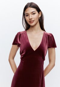 Robe en velours bordeaux avec un profond décolleté en V et des manches courtes à volants. Le tissu est doux, avec une texture lisse et une silhouette ajustée.
