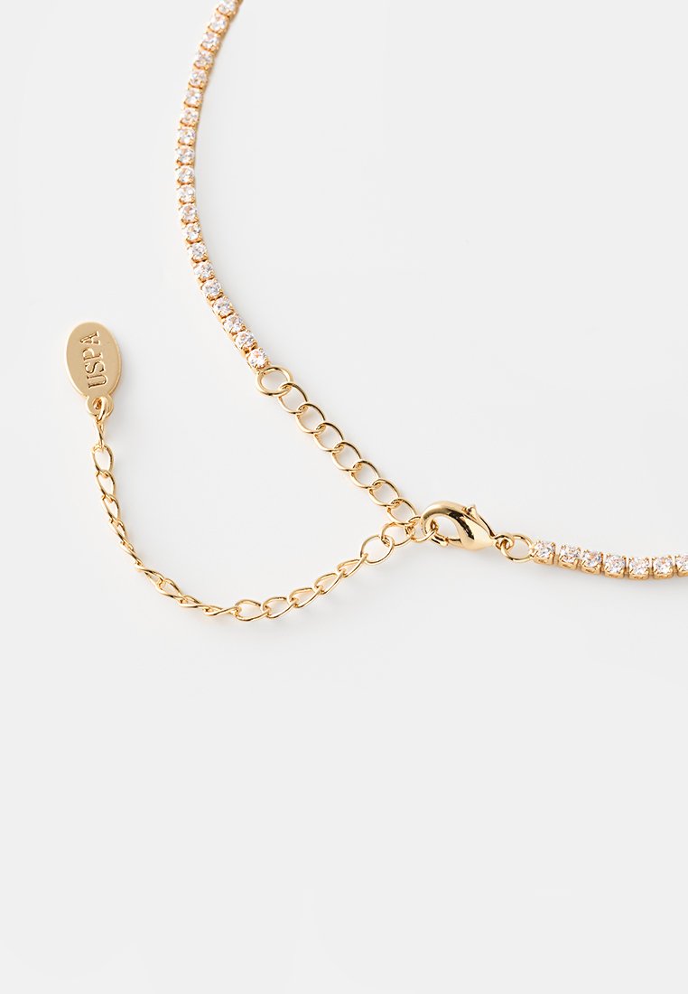 Collana in tono oro con una fila di cristalli trasparenti, dotata di chiusura a gancio e di una catena regolabile per la vestibilità. Etichetta incisa con "USPA."
