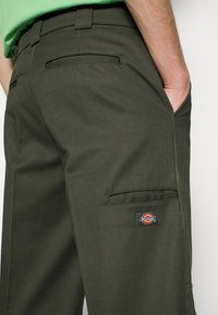 Mörkgröna arbetsbyxor gjorda av slitstark tyg, med en sidficka, två bakfickor och ett litet Dickies-märke.