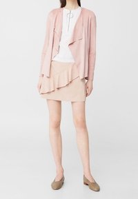 Giacca in suede rosa chiaro con design a strati e apertura frontale, abbinata a una gonna pastello con dettagli a ruches e ballerine beige.