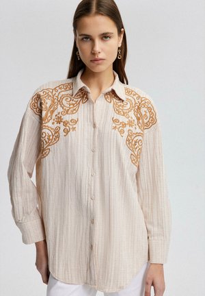 Camicia button-up di un beige chiaro con tessuto texturizzato. Presenta un intricato ricamo arancione sulle spalle con design floreali e a spirale. Maniche lunghe.