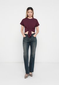 Bordeaux geknoopte crop top, korte mouwen, in combinatie met donkerblauwe flared jeans en lichtbruine puntige flats. Effen achtergrond.