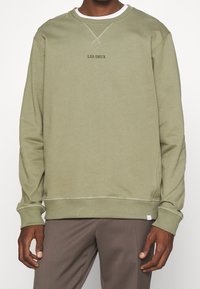 Uomo che indossa una felpa a girocollo di un leggero verde oliva con la scritta "LES DEUX" e pantaloni marroni, in piedi su uno sfondo semplice.