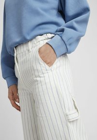 Sweatshirt bleu à manches longues associé à un pantalon blanc taille haute avec des rayures verticales bleues et des poches latérales.