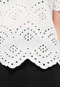 Λευκό κεντητό μπλουζάκι με κυματιστό τελείωμα, περίτεχνα σχέδια με κοψίματα και μαύρες λεπτομέρειες σε σκούρο φόντο.