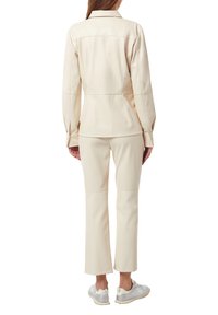 Ecrufarbene Kunstlederjacke mit Kragen, tailliertem Schnitt, langen Ärmeln und passenden ecrufarbenen Hosen. Details an den Schultern sichtbar.