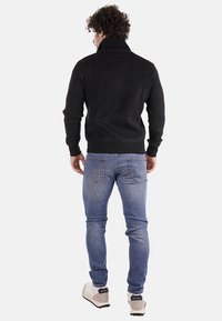 Maglione nero a coste con collo alto, indossato con jeans blu. Il maglione ha un design aderente e polsini aderenti, offrendo un aspetto texturizzato.